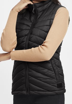 Frau trägt eine schwarze gesteppte ärmellose Weste über einem beigen, gerippten Langarmshirt, mit einer Hand berührt sie den Kragen der Weste.