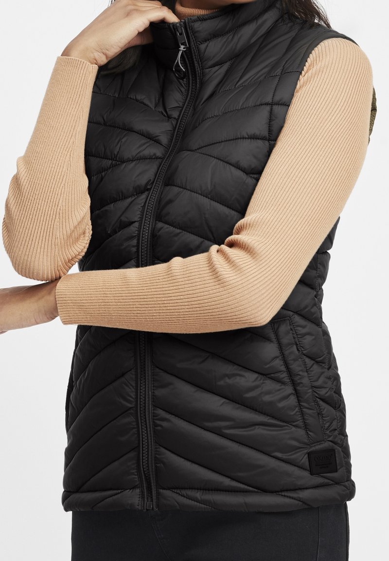 Femme portant une veste sans manches matelassée noire sur un haut beige à manches longues côtelé, une main touchant le col de la veste.