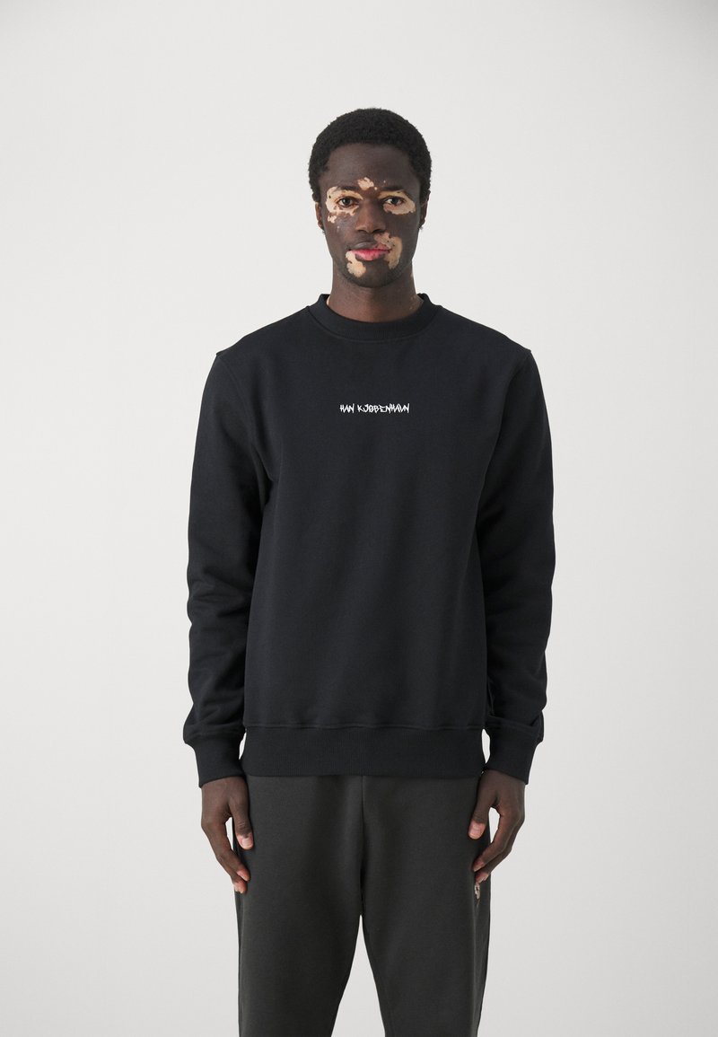 Black crew neck sweatshirt with white text "HW KUDLERMAN" on the chest; fabric appears to be cotton; model wearing gray pants.