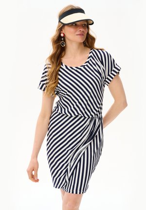 Kobieta w sukience w czarno-białe ukośne paski, czarno-beżowej daszku i dużych efektownych kolczykach, patrzy na bok z ręką na biodrze.