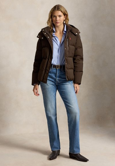 Femme portant une doudoune marron foncé sur une chemise bleu clair, un jean bleu, des bottes noires à bout pointu, debout devant un fond neutre.
