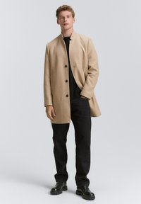 Beige wolmix jas met een staande kraag en vijf zwarte knopen. Draagt over een zwarte top, gecombineerd met zwarte broek en laarzen.