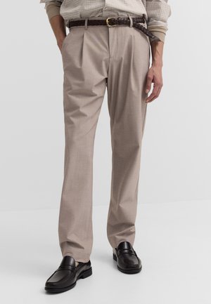 Mann trägt beige Anzugshosen mit vorderen Falten, braunen geflochtenen Gürtel, ein eingestecktes kariertes Hemd und schwarze Lederloafers.