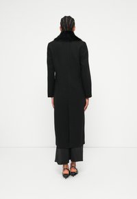 Cappotto lungo nero con colletto in pelliccia, presenta una linea dritta e maniche lunghe. La parte posteriore ha una cucitura centrale ed è abbinato a un outfit nero a strati.