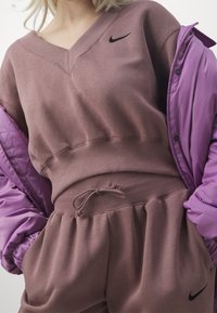 Hnědá mikina s výstřihem do V a žebrovaným lemem, kombinovaná s fialovou péřovou bundou. Má stahovací pás a logo Nike na hrudi.