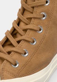 Brune semsket høy-top sneakers med sølvøyner og beige lisser, med en hvit gummisåle og sømdetaljer langs kantene.