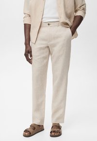 Homme portant un pantalon en lin beige, une chemise beige boutonnée sur un t-shirt blanc, et des sandales en daim marron à deux brides.