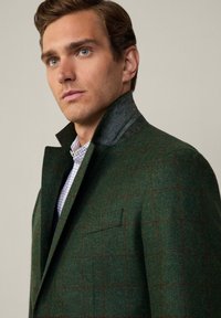 Grön herringbone-blazer med en strukturerad krage, med ett rutmönster i mörkröd. Tyget har en texturerad yta och skarpa slag.