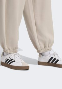 Biele tenisky s čiernymi pásikmi a béžovými podrážkami, nosené s joggerami béžovej farby s elastickými manžetami. Hladká textúra a športový dizajn.