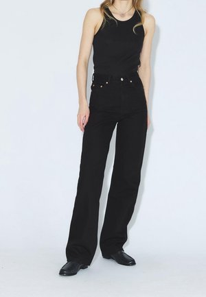 Femme portant un top noir sans manches, un pantalon noir taille haute à jambes larges et des chaussures noires, debout devant un fond blanc uni.