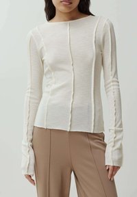Lichtbeige geribde langekous top met een wijde halslijn, verticale naden en uitlopende mouwen. Gecombineerd met strakke, op maat gemaakte bruine broeken.