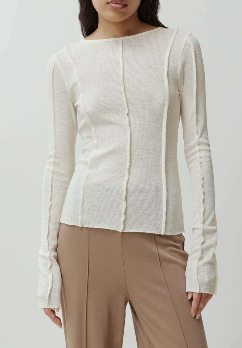 Lichtbeige geribde langekous top met een wijde halslijn, verticale naden en uitlopende mouwen. Gecombineerd met strakke, op maat gemaakte bruine broeken.