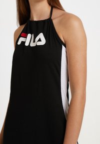Czarna sukienka na halter, posiadająca logo Fila w białym i czerwonym kolorze, z siateczkowymi białymi bokami oraz marszczonym dekoltem. Gładka tekstura materiału.