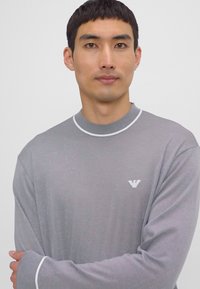 Maglione grigio a maniche lunghe con dettagli bianchi al colletto e ai polsini, caratterizzato da un piccolo logo ricamato sul petto. Tessuto morbido.