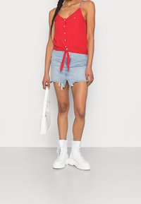 Top sin mangas rojo con cierre de botones y detalle de lazo en la cintura, combinado con una falda de mezclilla azul clara deshilachada y botas blancas con cordones.