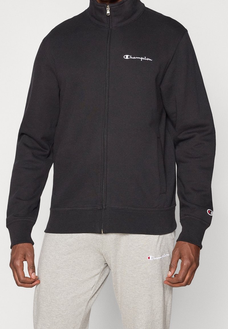 Sweat-shirt noir à fermeture éclair avec col montant, arborant un logo Champion blanc sur le côté gauche de la poitrine et des poignets et ourlet côtelés. Associé à un pantalon de survêtement gris.