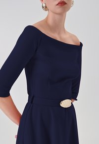 Dunkelblaues Kleid mit schulterfreiem Ausschnitt, Dreiviertelärmeln und einer strukturierten goldenen Gürtelschnalle als Akzent. Glattes Material.
