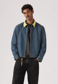 Άνδρας που φοράει μπλε καρό σακάκι με κίτρινο γιακά, μαύρο παντελόνι με μπρελόκ και μαύρη μπαντάνα γύρω από τον λαιμό, στέκεται με τα χέρια στις τσέπες.