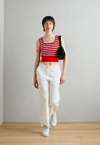 Crop top rayé rouge et blanc, pantalon large blanc et baskets blanches. Le mannequin tient un sac à main noir, debout devant un fond clair.