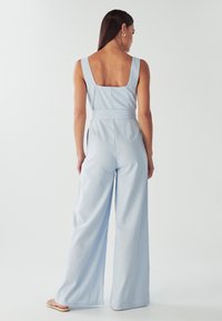 Salopette bleu clair avec un décolleté carré, jambes larges, taille ajustée et texture lisse. Présente une ouverture dans le dos et des accents minimalistes.