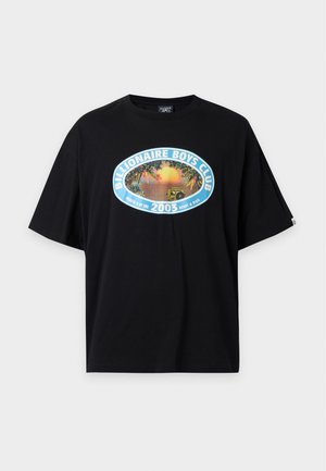 Mann steht mit einer Hand in der Tasche, trägt ein schwarzes Billionaire Boys Club T-Shirt, ausgewaschene schwarze Jeans und weiße Turnschuhe auf weißem Hintergrund.