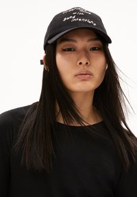 Schwarze Mütze mit weißem, gesticktem Text, mit einem gebogenen Schirm und einem Riemen an der Seite. Das Modell trägt ein schwarzes Langarmshirt.