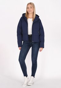 Marinblå puffjacka med huva, dragkedja fram, två sidfickor och quiltad textur, kombinerad med mörka jeans och vita sneakers.