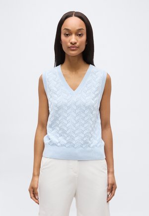 Femme portant un gilet en tricot sans manches bleu clair à col en V avec un motif texturé et un pantalon blanc, debout devant un fond uni.