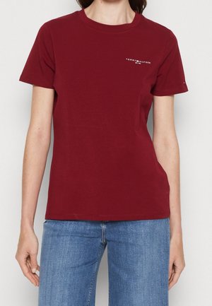 Femme portant un t-shirt uni rouge foncé à manches courtes avec un petit logo blanc Tommy Hilfiger sur la poitrine en haut à gauche et un jean bleu.