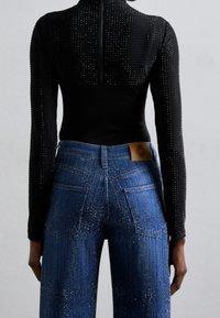 Henkilö, jolla on päällään musta pitkähihainen paita ja siniset farkut, joissa on kimaltavia koristeita, näkyy selkäpuolelta yksinkertaista taustaa vasten.