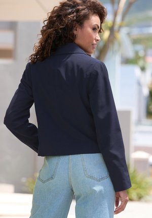 Veste cropped bleu marine avec un col structuré, des manches longues et une texture lisse, associée à un jeans léger taille haute.