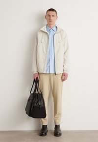 Lys beige zip-up jakke over en lys blå skjorte, kombinert med teksturerte beige bukser og svarte støvler. Holder en svart skulderveske.