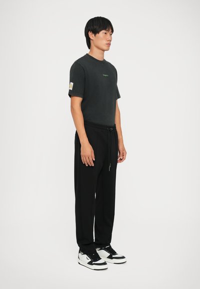 Sort bomulds T-shirt med grønt logo, parret med sorte joggers med snøre i livet og sneakers med hvide detaljer. Model står sidelæns.