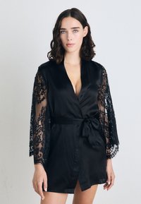 Robe di raso nero con lunghe maniche in pizzo e una vita con nastro. Il design presenta un profondo scollo a V e una parte anteriore leggermente aperta.