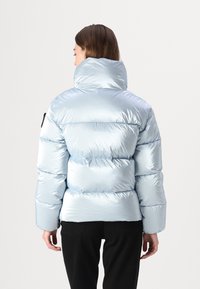 Gaisa jakas gaiši zilā krāsā, ar augstu apkakli, quilted dizainu, spīdīgu materiālu un melniem aksesuāriem uz kreisā piedurknes.