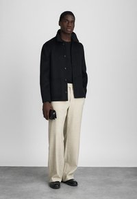 Muškarac u crnoj jakni na kopčanje, crnoj košulji, bež širokim hlačama, crnim cipelama, drži malu crnu torbicu, stoji na sivoj podlozi.