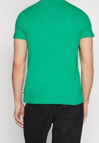 Zielona bawełniana koszulka, krótki rękaw, dopasowany krój, okrągły dekolt, gładka tekstura, akcent z logo przy dolnym brzegu, w zestawieniu z czarnymi spodniami.