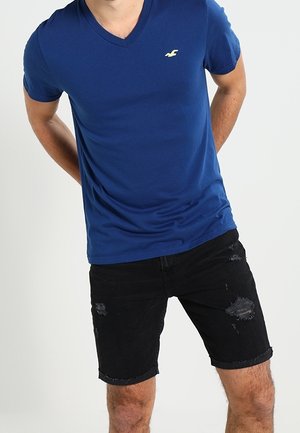 Niebieska koszulka v-neck z krótkimi rękawami, z małym żółtym logo, w połączeniu z czarnymi szortami dżinsowymi z przetarciami.