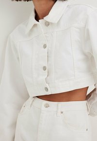 Veste en denim blanche coupée avec des boutons argentés, associée à un pantalon en denim blanc taille haute, montrant le ventre et des poches.