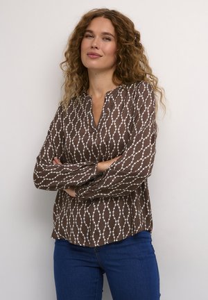 Brown long-sleeve blouse featuring a pattern of white dots and wavy lines, with a notch neckline and relaxed fit. Paired with blue jeans.