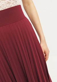Donna che indossa una gonna bordeaux a vita alta e plissettata e un top di pizzo chiaro, in piedi con il braccio destro rilassato al suo fianco.