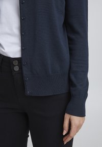 Marinblå stickad cardigan med knappstängning framtill, ribbade mudd och fåll. Åtföljs av svarta tajta jeans. Närbild på ärm och midjeområde.