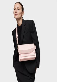 Rosa Leder Schultertasche mit gefaltetem Design und geprägt Logo, ausgestattet mit einem dicken Tragegriff und geschichteten Fächern. Getragen zu einem schwarzen Blazer.