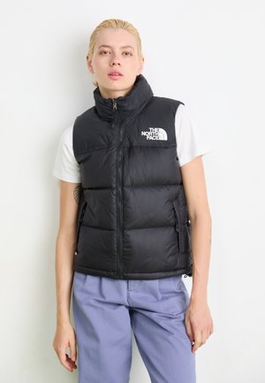 Zwarte puffervest met hoge kraag, voorrits en logo op de linkerborst. Gepaard met lichtpaarse broeken en een wit T-shirt.