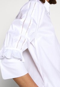 Camicia bianca con maniche voluminose, caratterizzata da polsini con volant e dettagli ricamati lungo le cuciture delle maniche. Tessuto liscio e leggero.