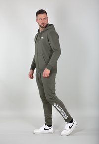 Felpa con zip color verde oliva, dotata di tasche frontali, abbinata a pantaloni jogger coordinati con stampa del logo lungo la gamba sinistra. Sneakers bianche completano il look.