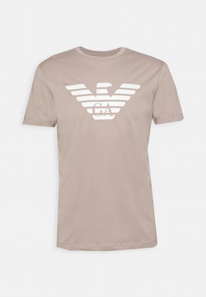 Beżowy t-shirt z krótkim rękawem z białym stylizowanym logo orła i inicjałami GA na środku klatki piersiowej.