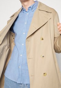 Vaaleanbeige kaksirivinen trench-coat, jossa on suuret napit, päällä vaaleansininen kauluspaita. Näkyvät yksityiskohdat sisältävät läpät ja taskut.