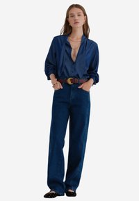 Blauwe denim shirt met een losse pasvorm en lange mouwen, gecombineerd met hoge blauwe jeans. Afgemaakt met een leren bruine riem en zwarte schoenen.