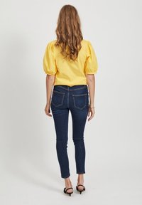Żółta bluzka z bufiastymi rękawami w połączeniu z ciemnoniebieskimi wąskimi dżinsami, z tylnymi kieszeniami i gładką teksturą denimu. Całość dopełniają proste czarne szpilki.